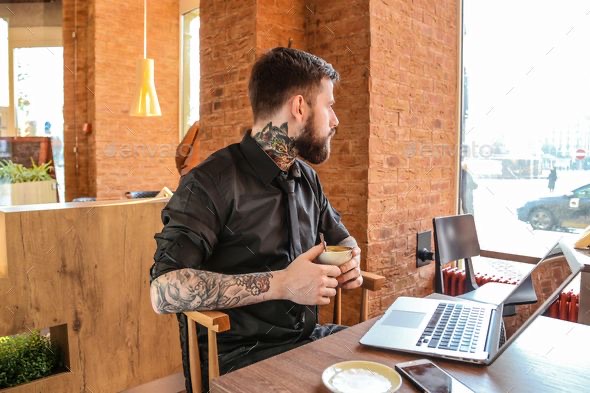 A man in a black shirt sits at a cafe table with a laptop and a cup of coffee, looking out of the window