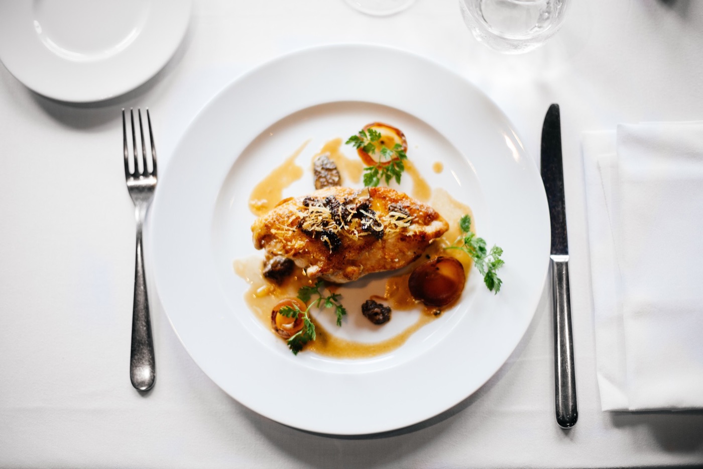 Overhead view of a plated fine-dining chicken dish with morel mushrooms, roasted shallots, and herb garnish on a white tablecloth