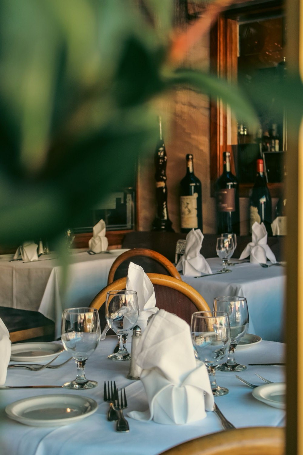 Restaurant tables laid with folded napkins and wine glasses, with wine bottles behind